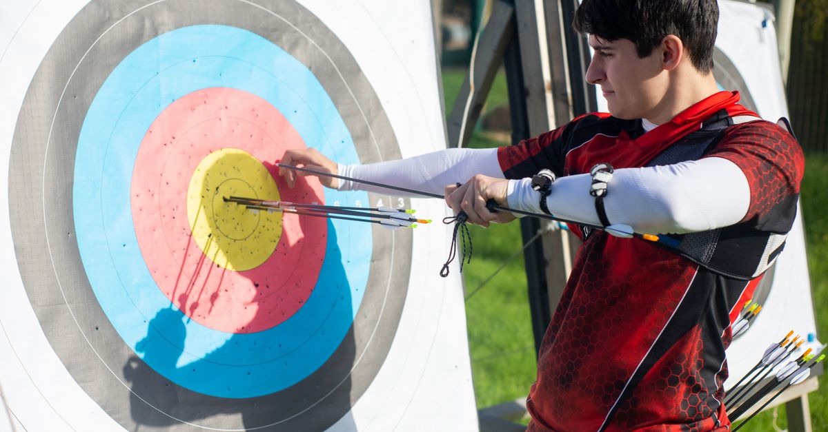 Focus on man aiming with bow and arrows at target during outdoor archery practice.